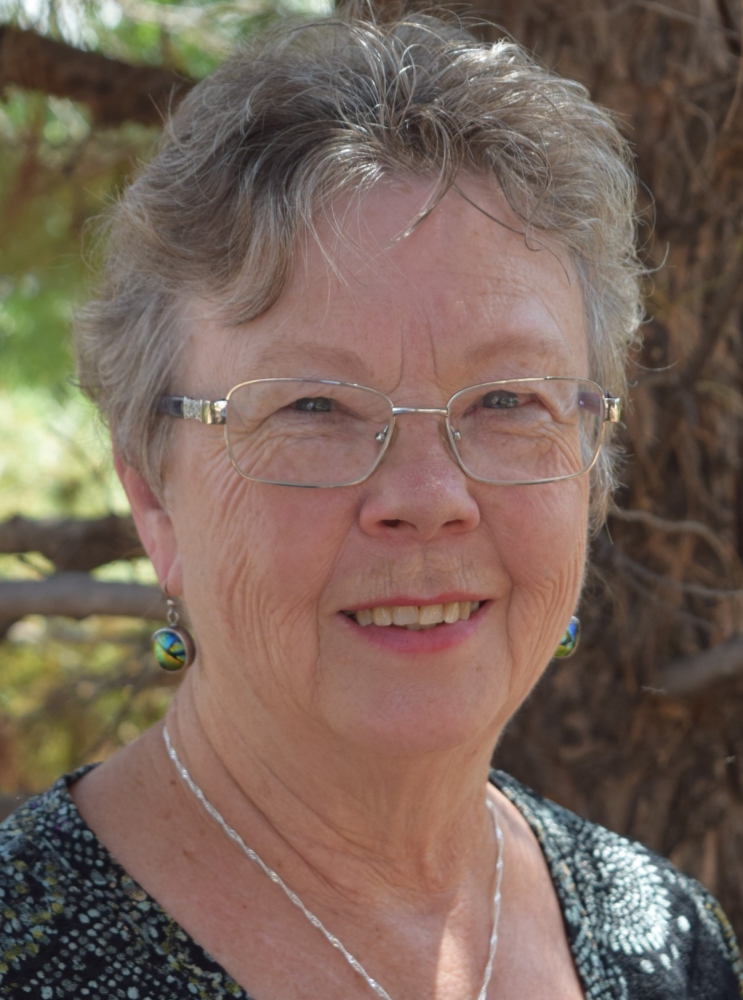 Becky Cerling Powers is an older lady with short, curling, graying hair, small slanted blue eyes and silver-rimmed glasses. She is wearing a black, blue, gold and green patterned top, round drop earrings that match colors with her top, and a silver chain around her neck. She is smiling, and smile wrinkles radiate downward from her eyes. The picture shows only her face and shoulders.