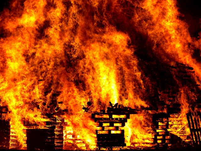 A fire is destroying a building. Bright orange and yellow flames glow through holes in all that is left of the lower structure. A great tower of flame mingled with black smoke rises above and fills the entire picture frame. 